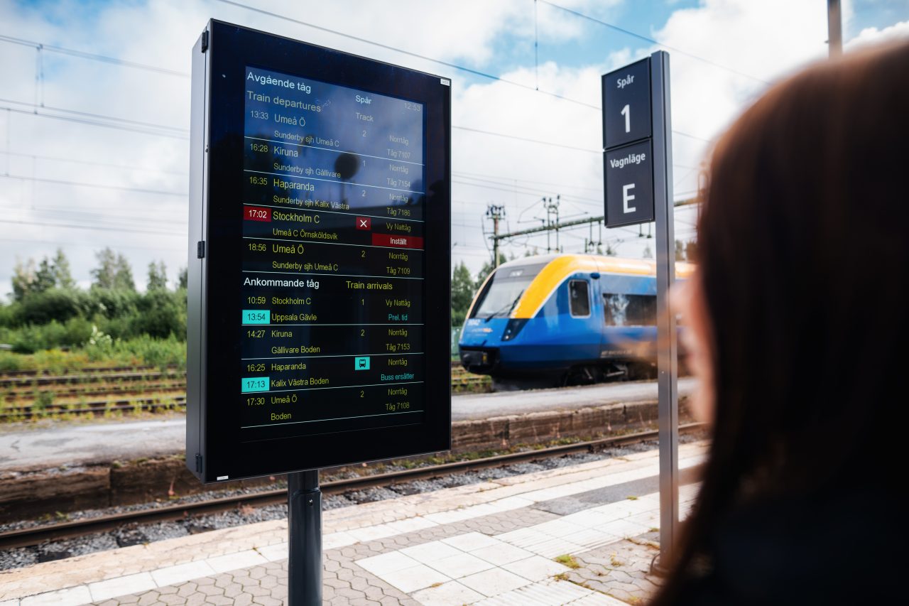 Person tittar på informationstavla vid tågstation. I bakgrunden står ett Regina X52-fordon.