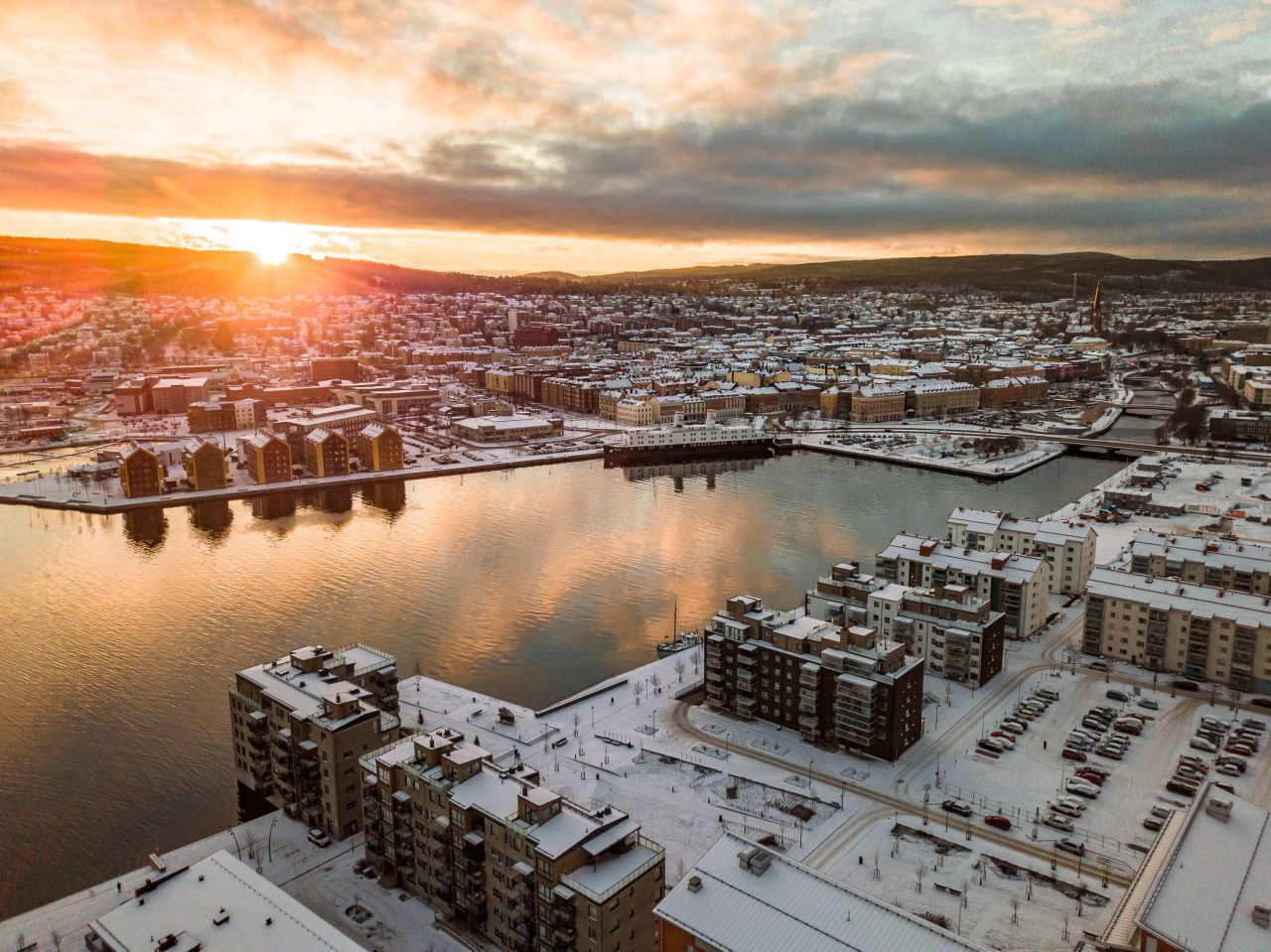 Flygfoto över ett vinterklätt Sundsvall i solnedgång, med snötäckta byggnader och vattnet som speglar himlens färger.